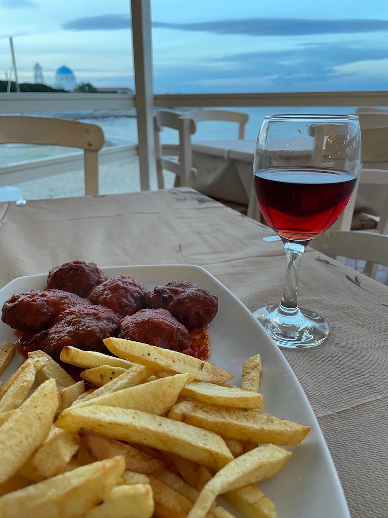 A meal of keftedes and patates with a glass of red wine, the sea in the background.